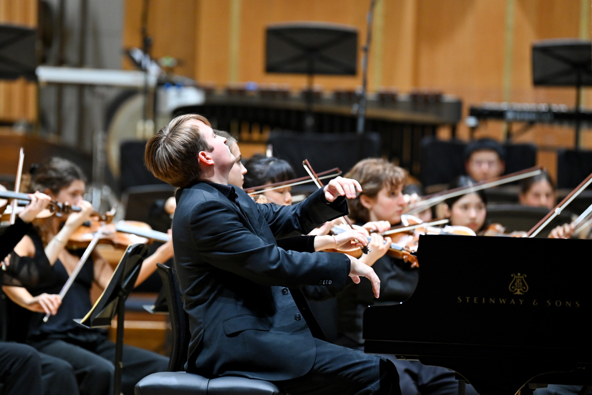 A student wearing smart black attire, playing the piano in an orchestra, with an orchestra being conducted in the background.